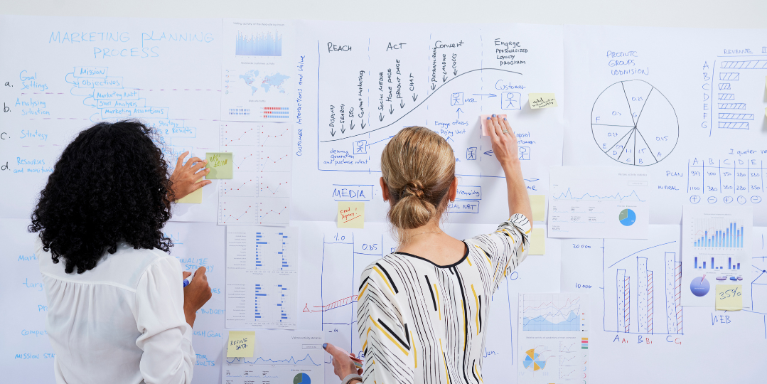 Two women planning a marketing strategy in front of a whiteboard covered with charts, graphs, and sticky notes, illustrating the importance of consistent website oversight as part of a WordPress maintenance plan.