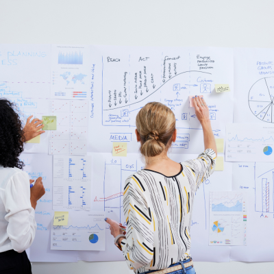Two women planning a marketing strategy in front of a whiteboard covered with charts, graphs, and sticky notes, illustrating the importance of consistent website oversight as part of a WordPress maintenance plan.